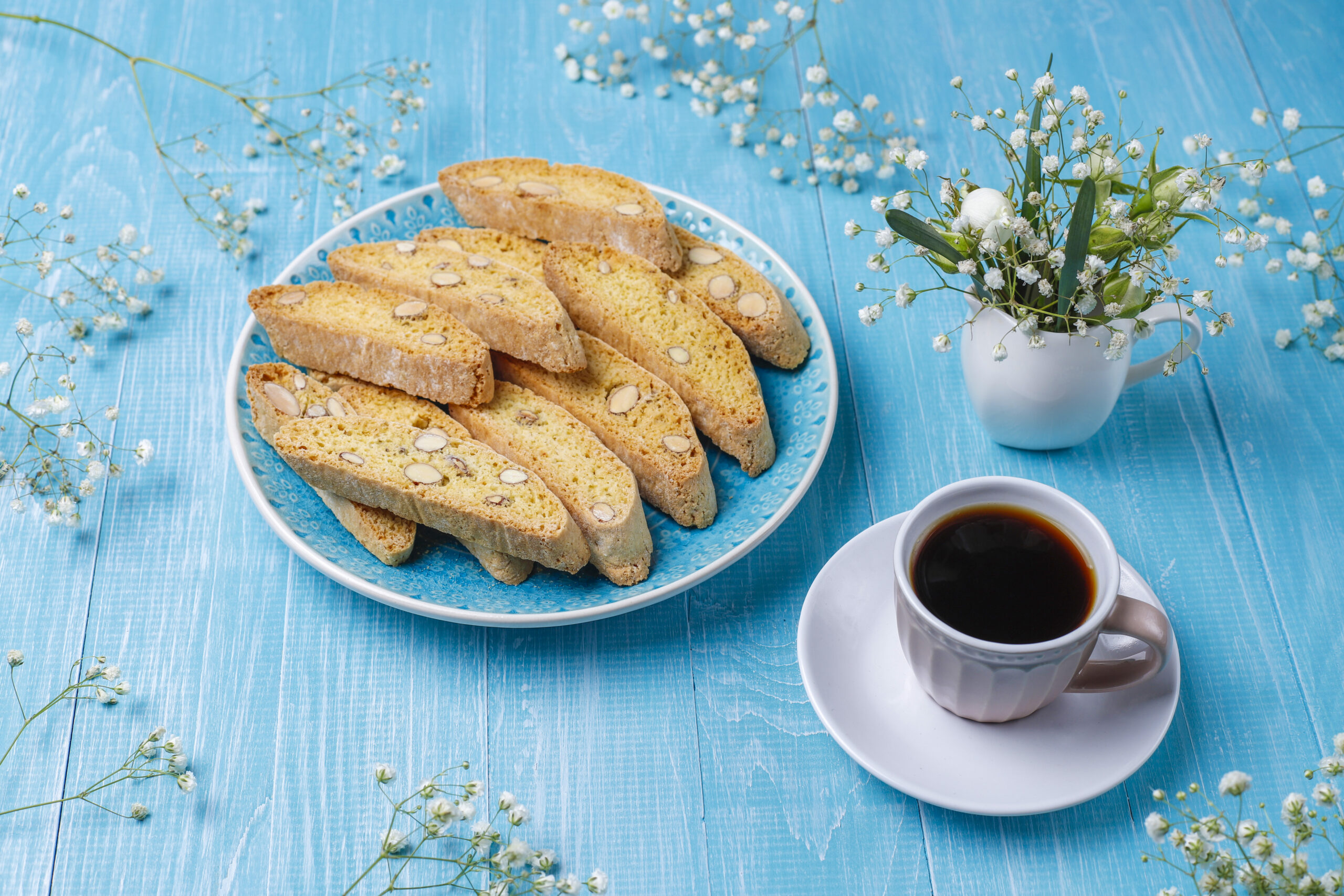 A plate of biscotti and a cup of coffee on a blue wooden table, surrounded by delicate flowers.